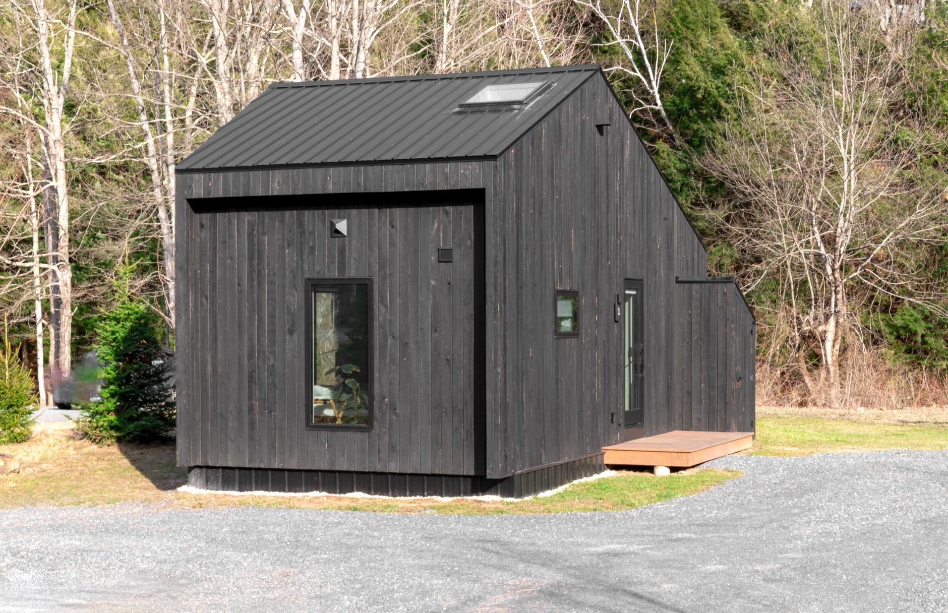 Small wooden tiny house with a sleeping loft window and a family-sized porch in a wooded lot